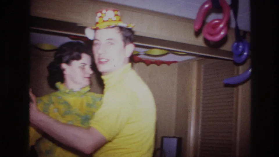 A man in a yellow shirt and party hat smiles toward the camera while dancing with a woman in a yellow patterned dress.