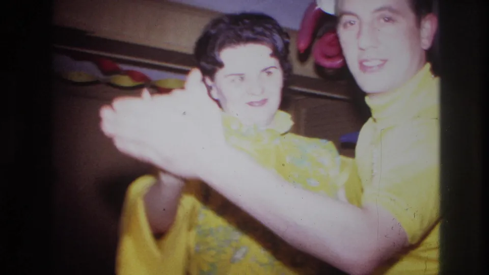 A close 8mm frame shows the couple dancing with clasped hands, their yellow outfits bright under indoor party lighting.