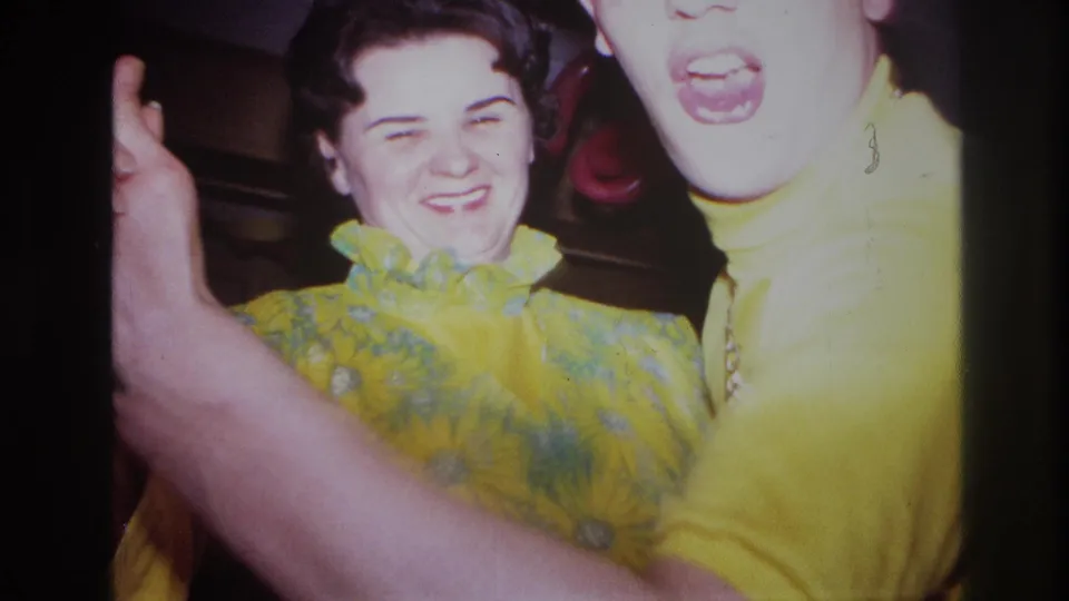 In 1967 Ottawa home-movie footage, two adults in bright yellow outfits laugh and dance near the camera inside a decorated room.