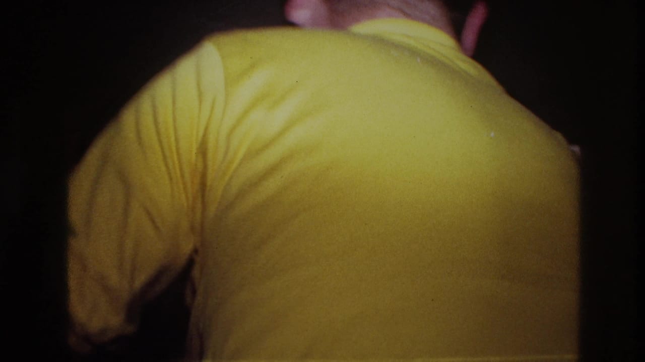 A man in a yellow shirt is seen from behind in a dim indoor room.