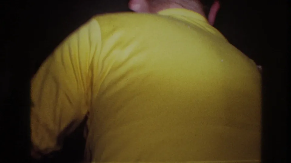 A man in a yellow shirt turns away from the camera during the Ottawa indoor party scene, with the room mostly dark around him.