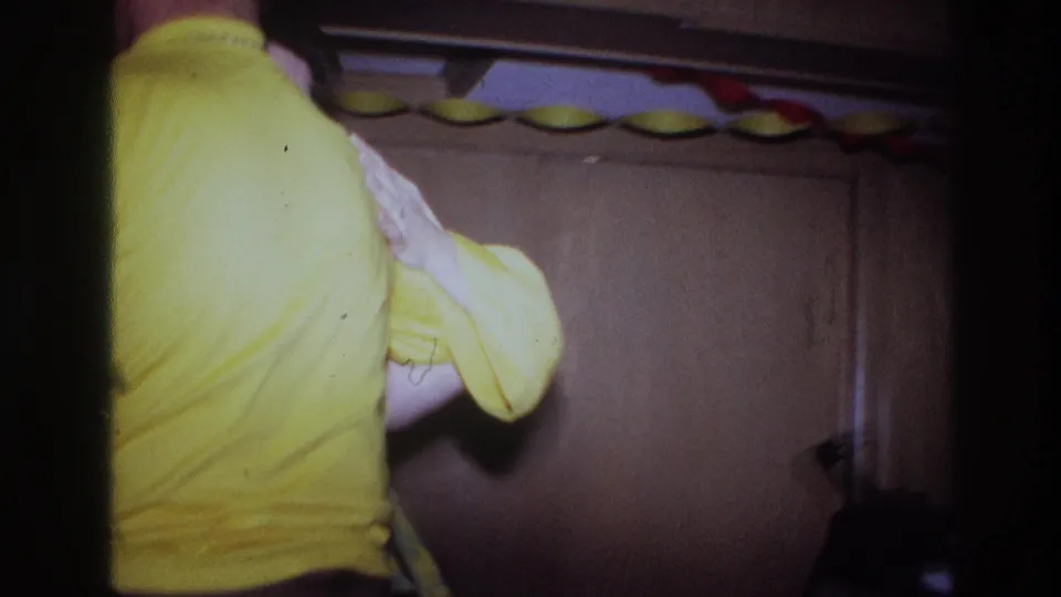 A close 8mm frame shows the pair from behind as they continue dancing near wood-paneled doors and paper decorations.