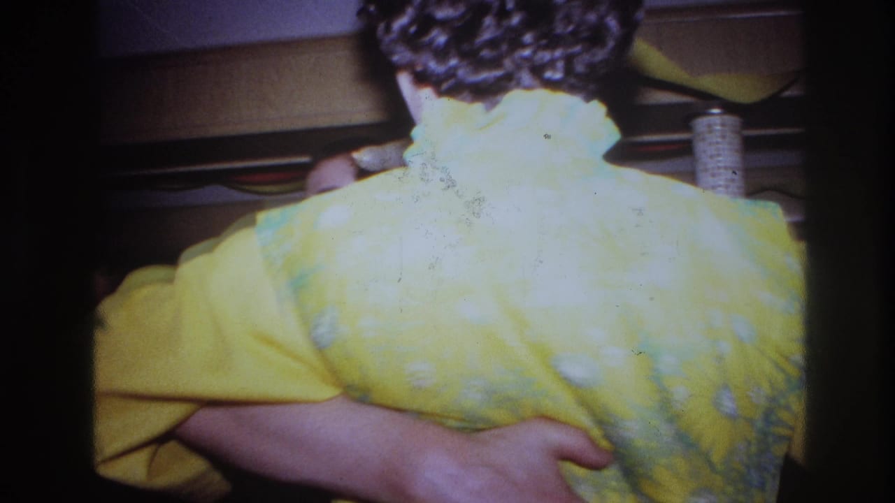1967 Ottawa home-movie frame showing a woman in a bright yellow floral dress held by a dance partner in a dim decorated interior.
