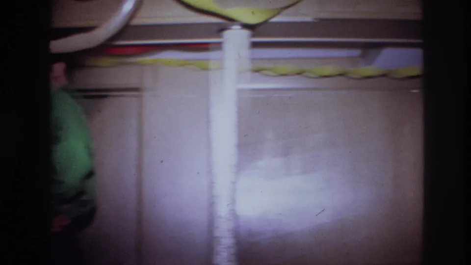 1967 Ottawa home-movie frame showing a partially framed man in green beside a support pole, wood paneling, and party streamers.