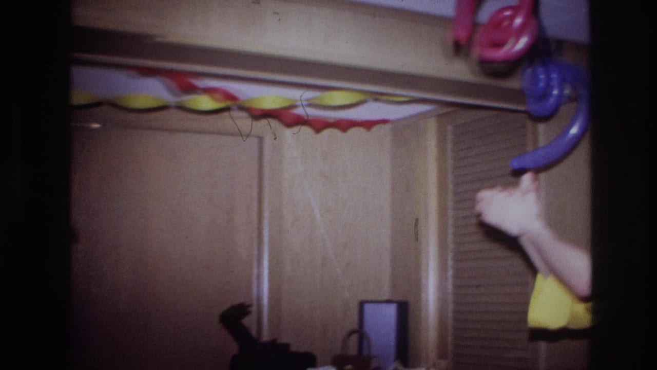 Ottawa 1967 home-movie frame showing a private party room with paper garland, hanging balloons, wood doors, and a cropped adult in yellow.
