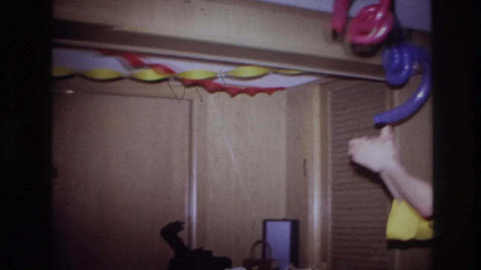 Ottawa 1967 home-movie frame showing a private party room with paper garland, hanging balloons, wood doors, and a cropped adult in yellow.