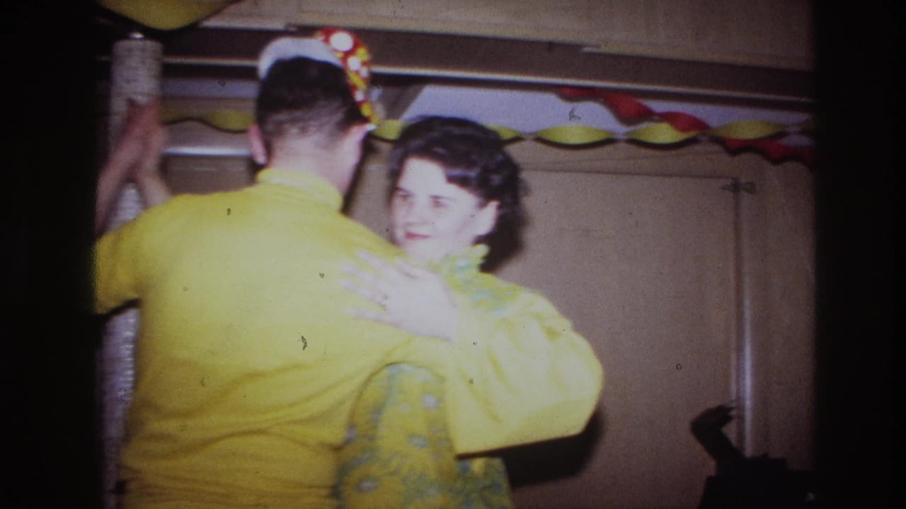 In a 1967 Ottawa home-movie frame, an adult couple in yellow dance beneath paper garlands in a wood-paneled interior.