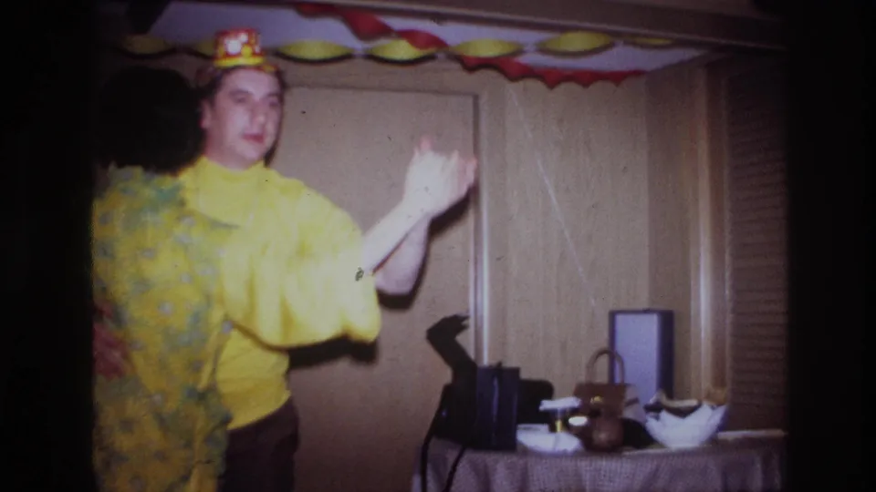 The man in a yellow shirt and polka-dot party hat dances near a small table in the 1967 Ottawa interior scene.