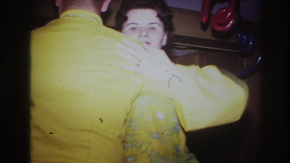 1967 Ottawa home-movie frame of a woman facing the camera as she dances with a man in a yellow shirt inside a decorated room.