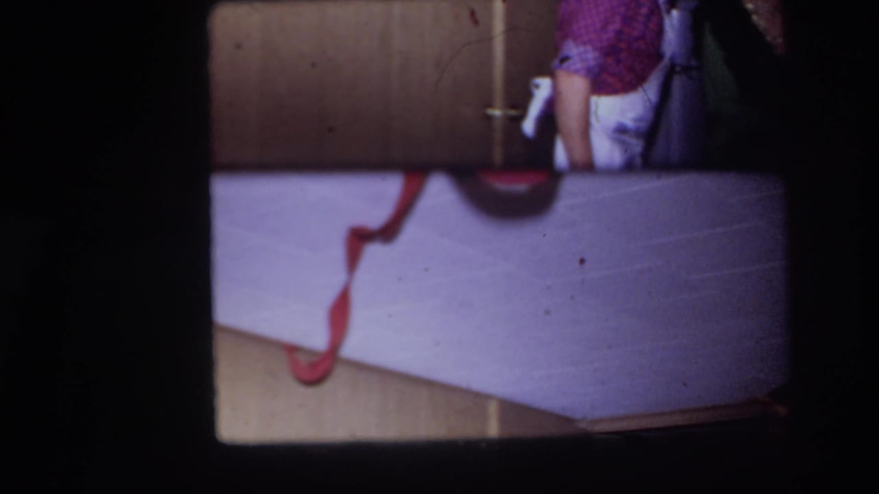 1967 Ottawa home-movie frame showing a cropped person in a checkered shirt beside wood cabinet doors and a pale surface with a red cord.