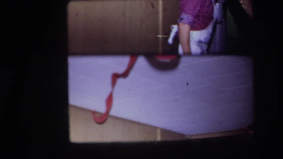 1967 Ottawa home-movie frame showing a cropped person in a checkered shirt beside wood cabinet doors and a pale surface with a red cord.
