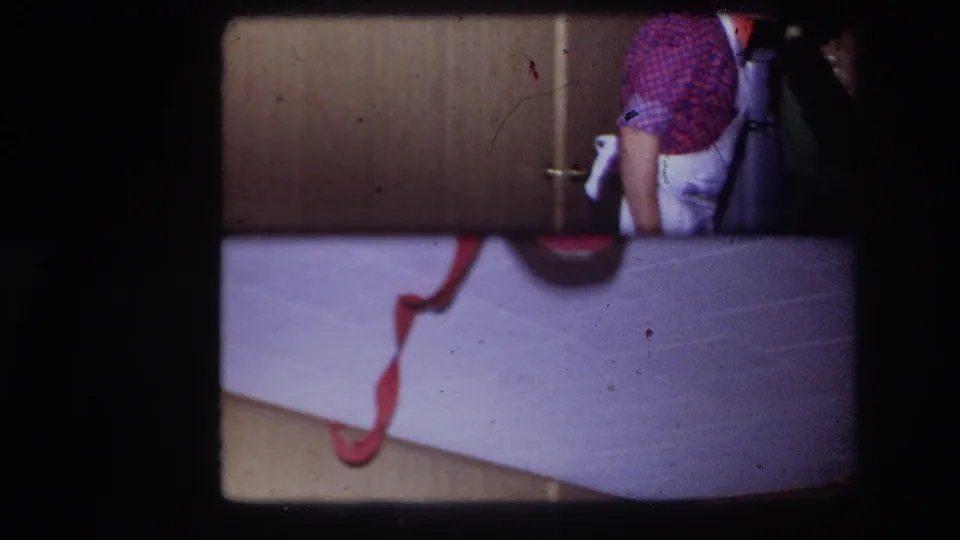 1967 Ottawa 8mm frame of a person in a checkered shirt and white pants standing near wood cabinetry, partly obscured by the film frame.