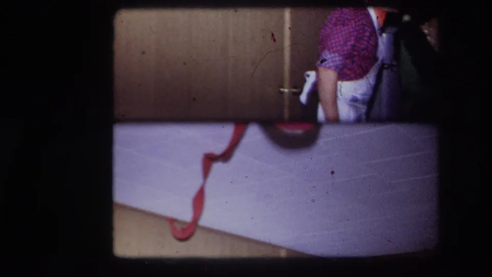 1967 Ottawa home-movie frame with a cropped person at right, wood cabinet doors behind, and a red cord across a pale foreground surface.