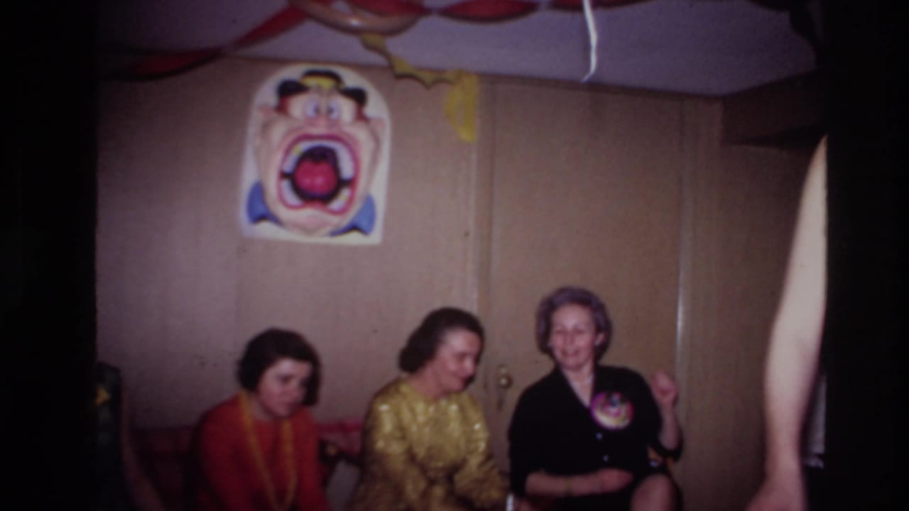Three women sit on a couch below a clown face poster in a party room.