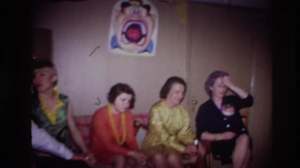 Adults sit together in the Ottawa party room, with bright dresses, yellow leis, a clown poster, and a table lamp visible.