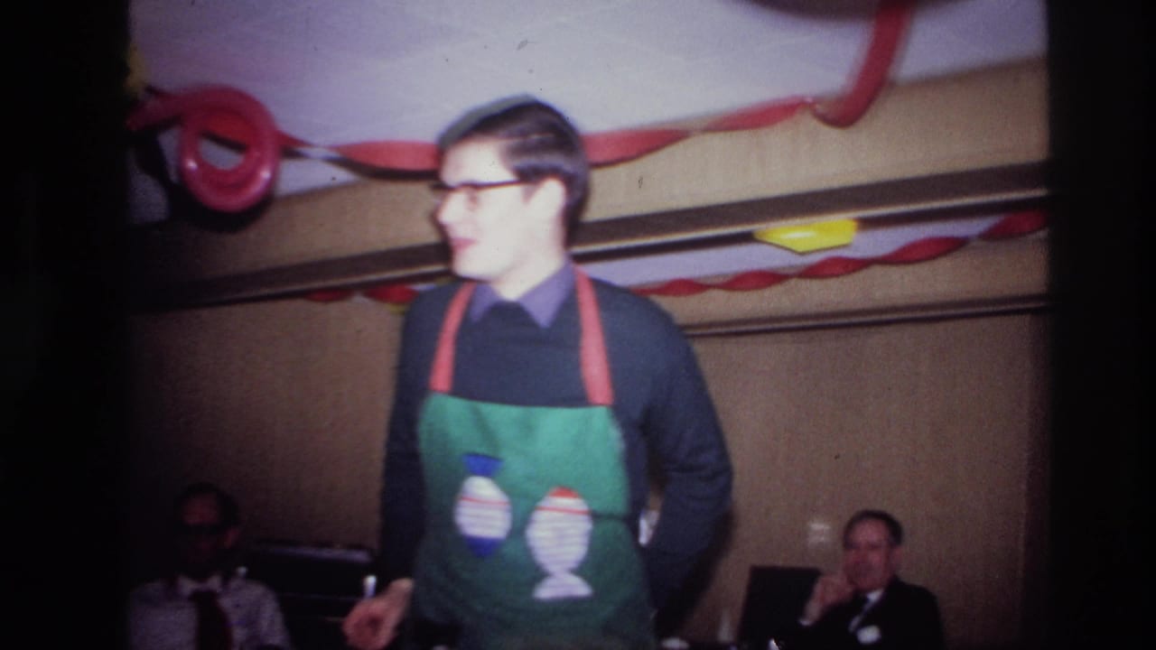 In Ottawa 1967 home-movie footage, a man in glasses and a green apron stands beneath streamers during an indoor party.