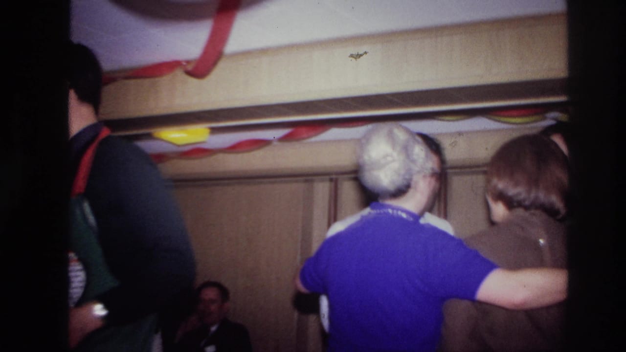 Two adult guests stand arm in arm beneath party streamers in a private Ottawa interior, with seated guests behind them.