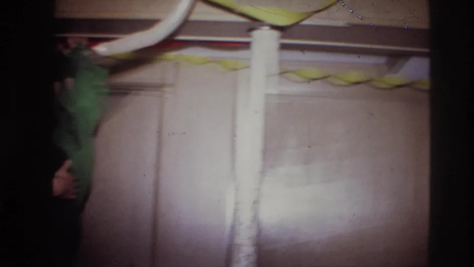 A handheld 8mm frame catches motion blur, a green apron at the edge, balloons overhead, and the light-colored wall of the party room.