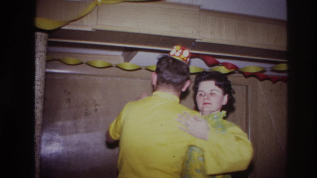 The man, wearing a polka-dot party hat, faces away while dancing with a woman in a yellow patterned dress indoors.