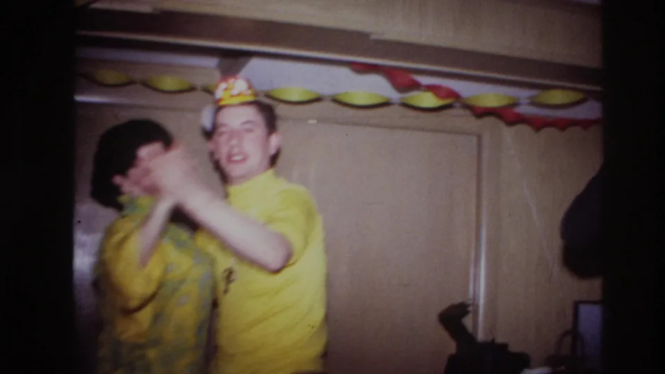 A close 8mm frame shows the dancing couple turning, with the man in a party hat and yellow shirt near the camera.