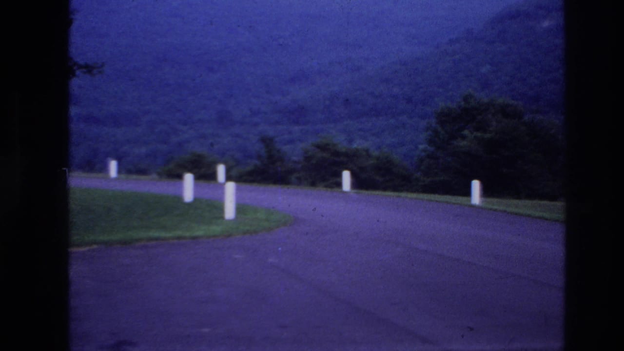Winding paved road with white roadside posts, grass verge, trees, and wooded hills in the background.
