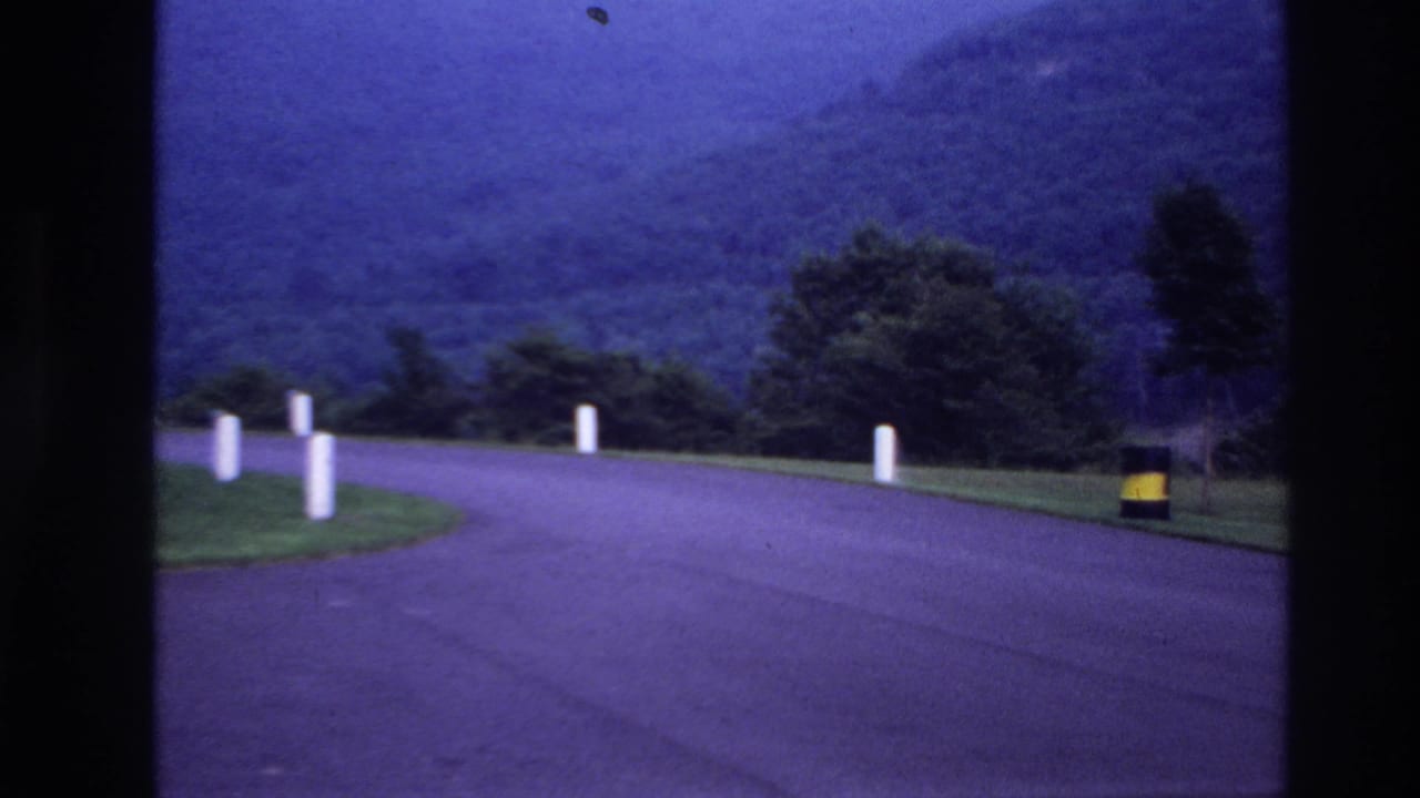 Blue-tinted 1967 Ottawa-area frame of a quiet bend in the road with roadside posts, trees, hills, and a yellow-and-black barrel.