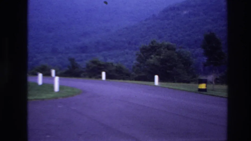 Blue-tinted 1967 Ottawa-area frame of a quiet bend in the road with roadside posts, trees, hills, and a yellow-and-black barrel.
