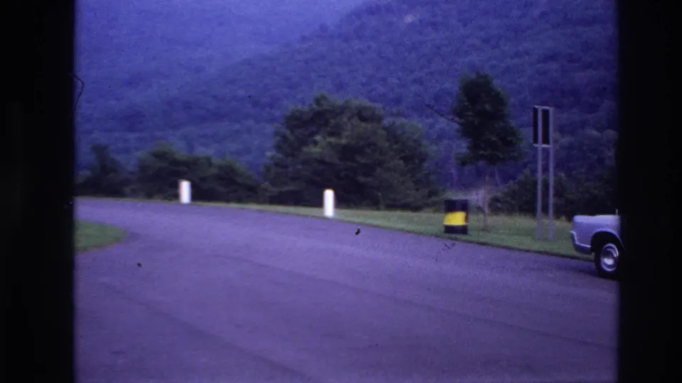 1967 Ottawa-area home-movie frame showing a wooded roadside curve, white posts, signs, a barrel, and the rear of a blue car.