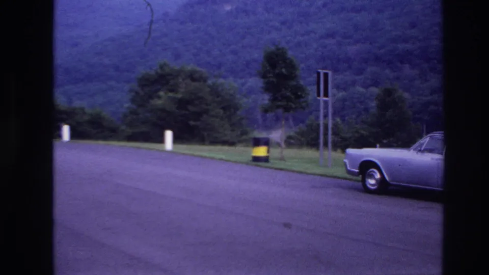 1967 Ottawa-area frame with a blue car partly visible beside a curved road, roadside signs, barrel, trees, and distant wooded hillside.