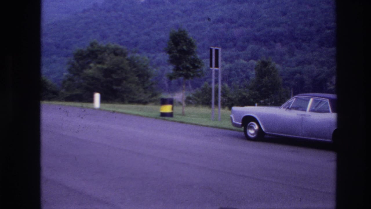 1967 Ottawa home-movie frame showing the front portion of a silver sedan beside a grassy roadside area and wooded hillside.