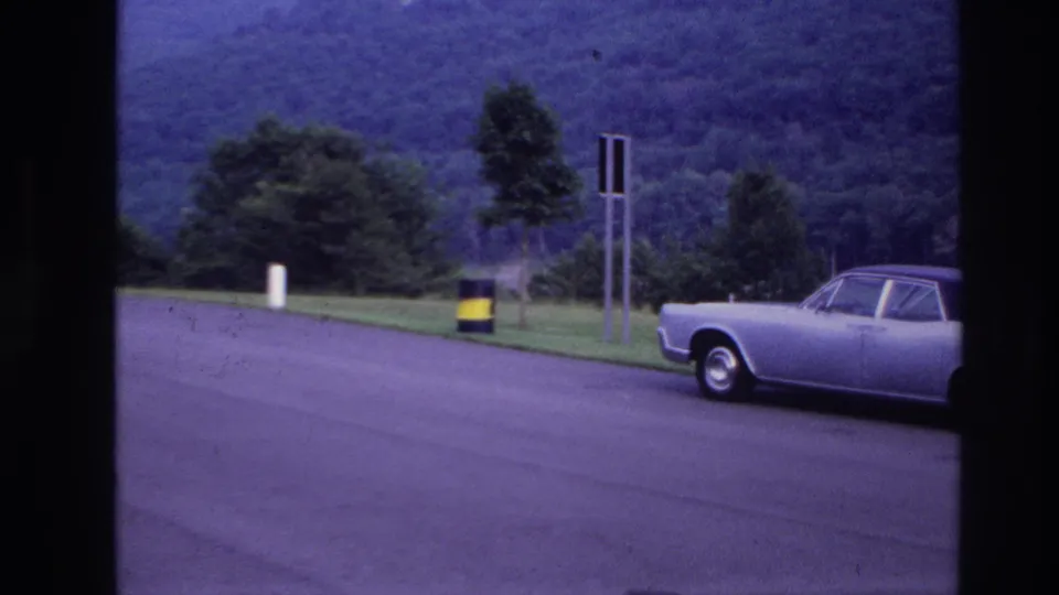 1967 Ottawa home-movie frame showing the front portion of a silver sedan beside a grassy roadside area and wooded hillside.