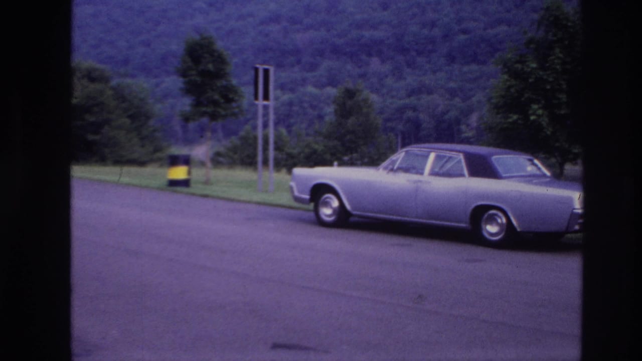 Silver sedan parked near grass with a striped barrel, signs, trees, and wooded hill in the background.
