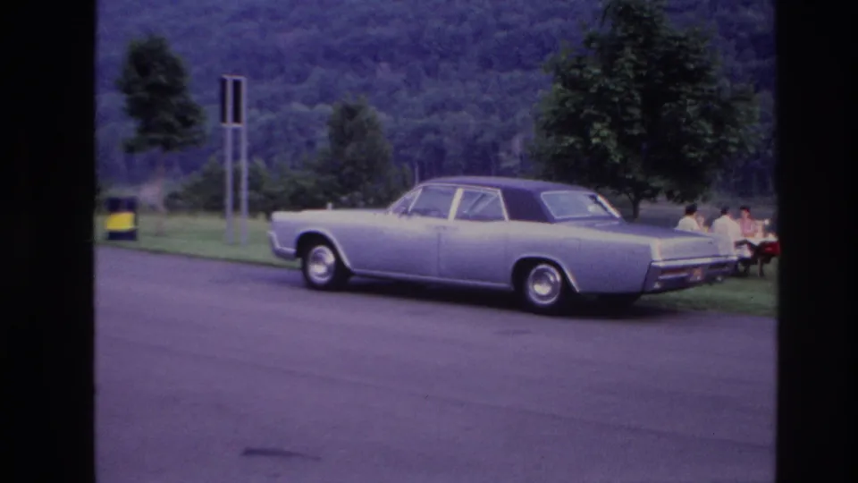 A 1967 Ottawa roadside picnic scene begins to emerge, with the parked sedan in front and people seated on the grass at right.