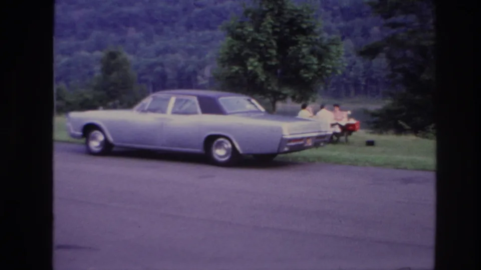 1967 Ottawa home-movie frame showing a pale sedan near a lakeside picnic table with seated family members in the background.