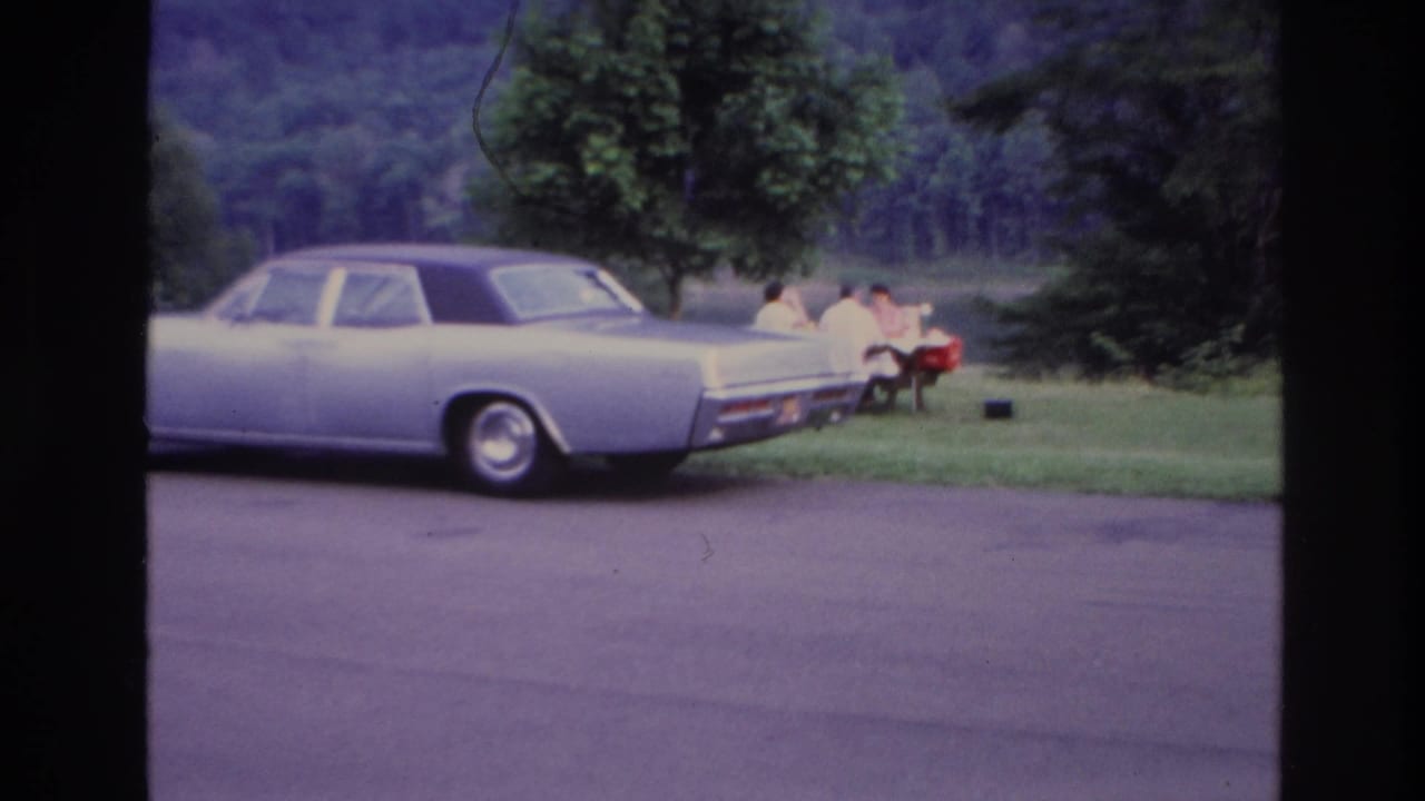 A 1967 Ottawa lakeside picnic scene with a pale sedan in the foreground and family members seated near a red cooler.