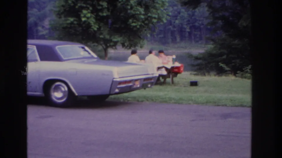 Closer 1967 Ottawa home-movie view of the sedan's rear and a family picnic table set on grass by the water.