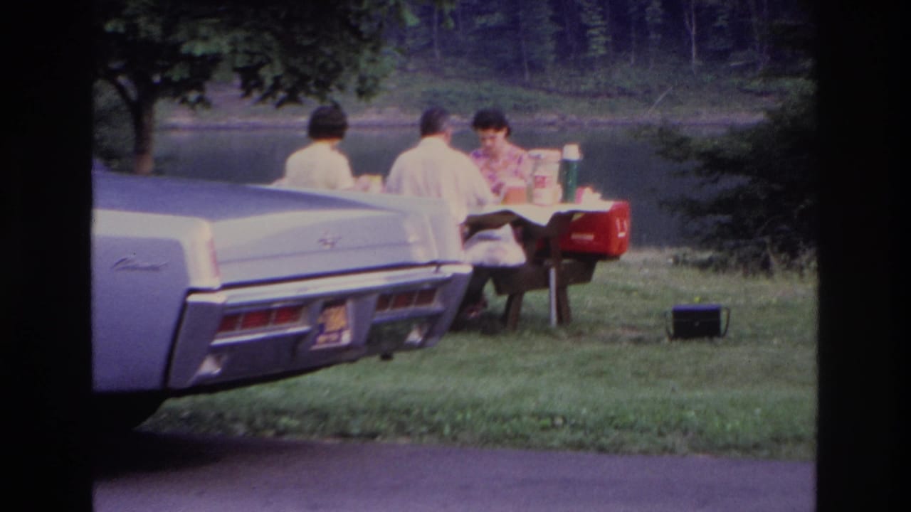 1967 Ottawa home-movie frame of adults at a picnic table near a lake, with a pale classic car in the foreground and a red cooler nearby.