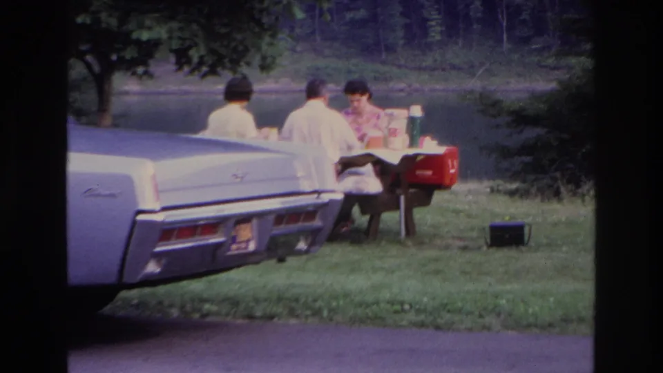 1967 Ottawa home-movie frame of adults at a picnic table near a lake, with a pale classic car in the foreground and a red cooler nearby.