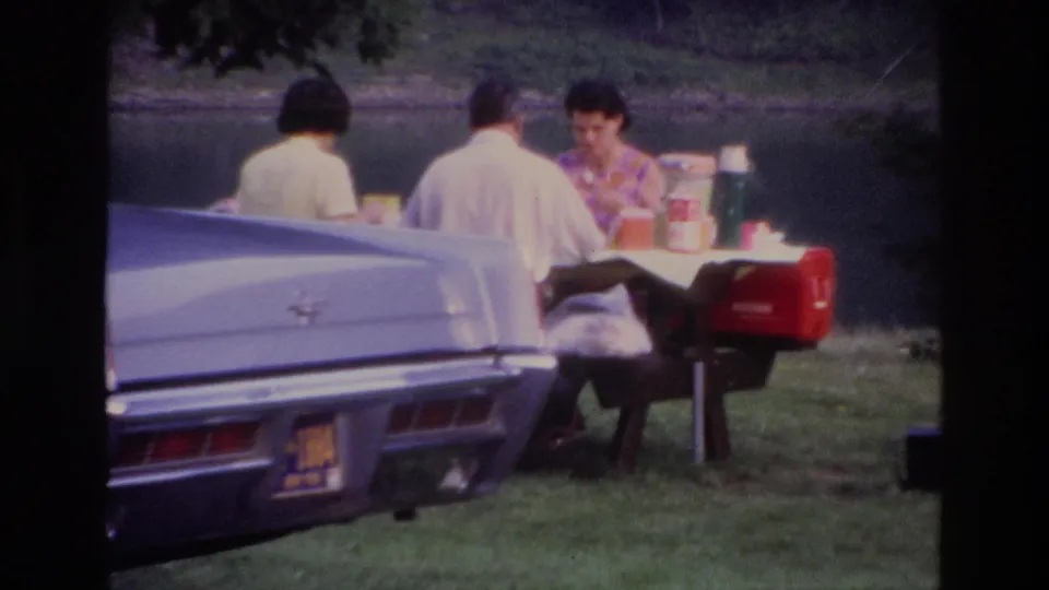 Ottawa 1967 lakeside picnic frame showing three adults at a table, with the rear of a pale classic car prominent in the foreground.