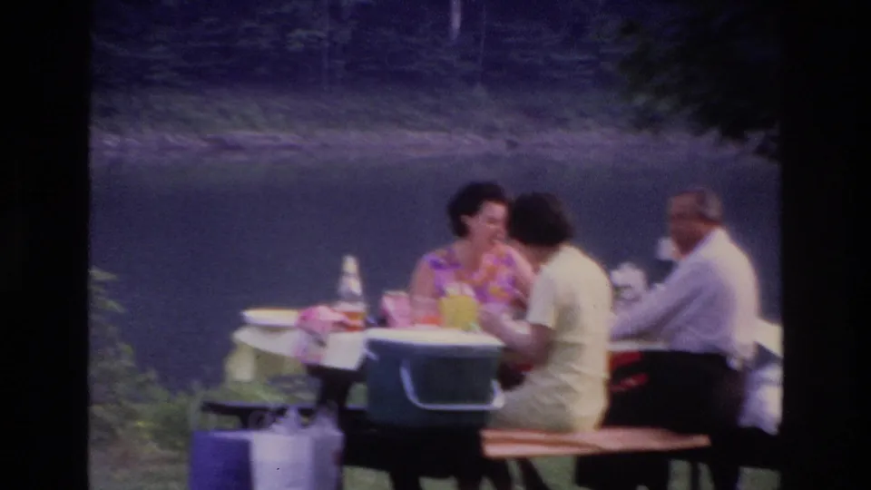 Closer 1967 Ottawa picnic view of adults seated by lake water, with table items, a cooler, bottles, and a bench visible in the foreground.