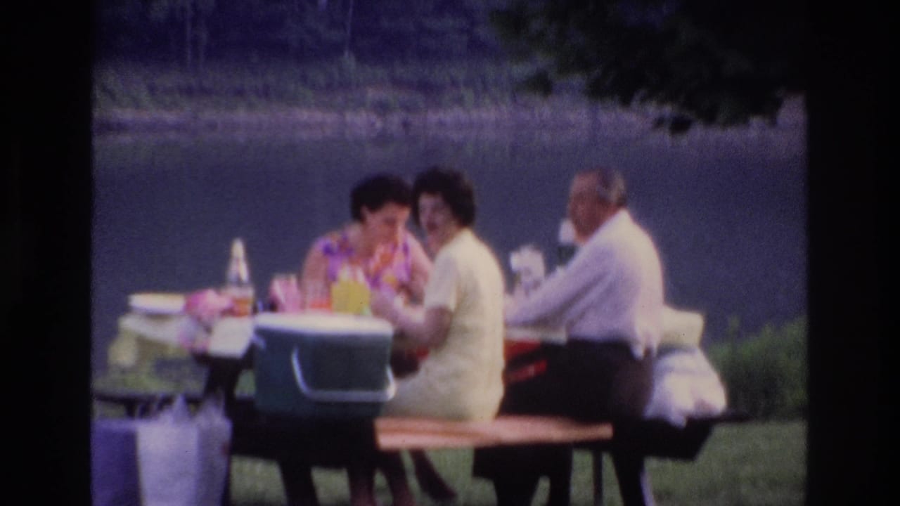 1967 Ottawa home-movie frame of three adults seated at a park picnic table by the water, with food, bottles, and a green cooler visible.