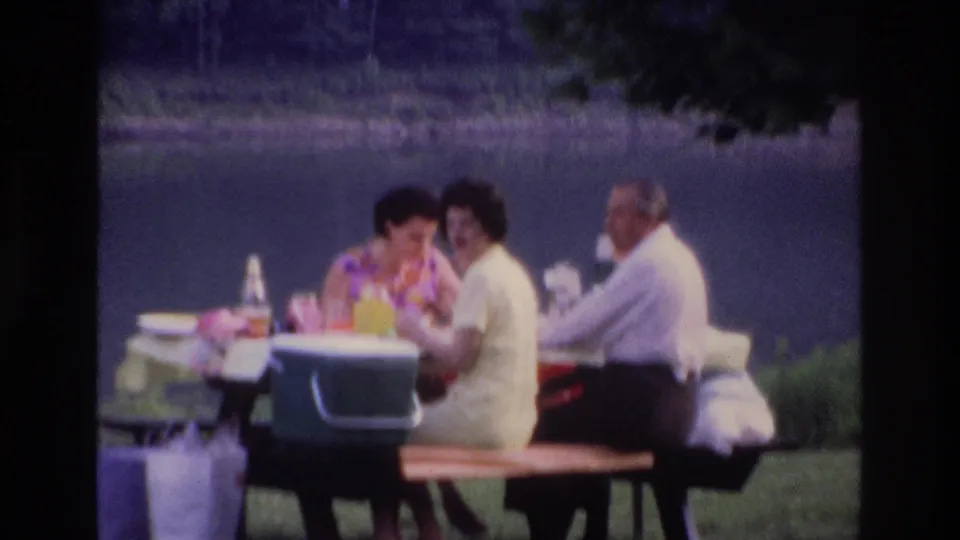 1967 Ottawa home-movie frame of three adults seated at a park picnic table by the water, with food, bottles, and a green cooler visible.