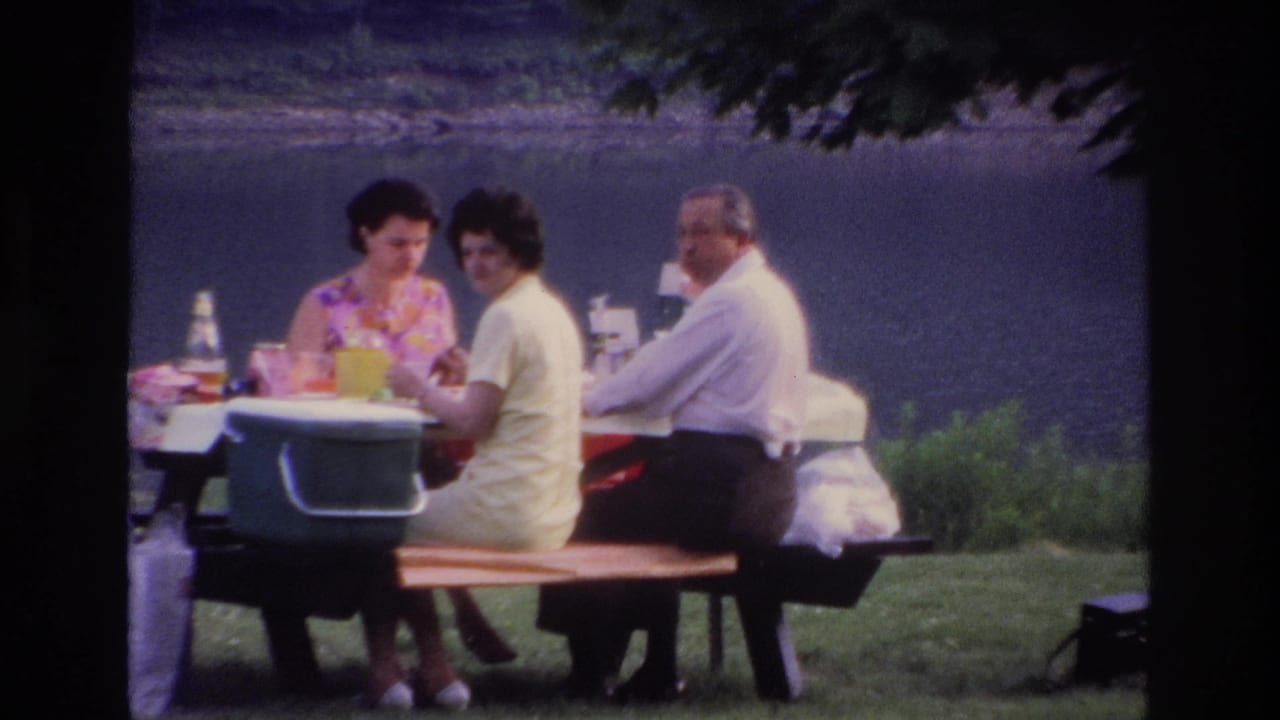1967 Ottawa frame showing two women and a man at a picnic table near a lake, with bottles, food, and a green cooler on the table.