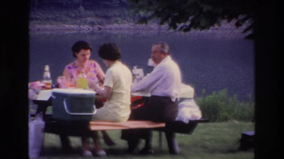 1967 Ottawa home-movie still of three adults at a lakeside picnic table, with a cooler and tableware visible under tree shade.