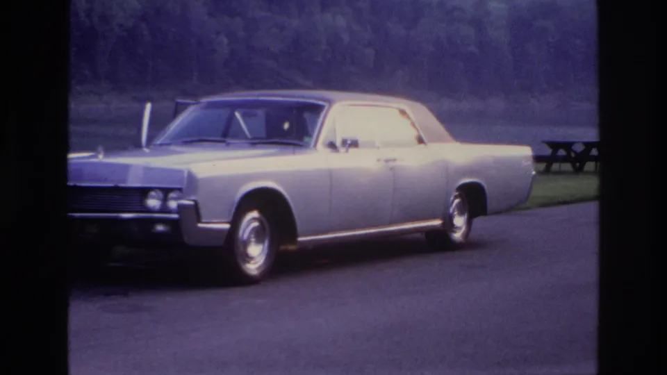 Wider Ottawa 1967 frame of the parked silver sedan near a grass verge, picnic table, water, and forested hillside.