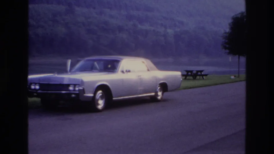 Wide 1967 Ottawa home-movie shot of the silver sedan parked near a lakeside or riverside road with picnic table and tree line.