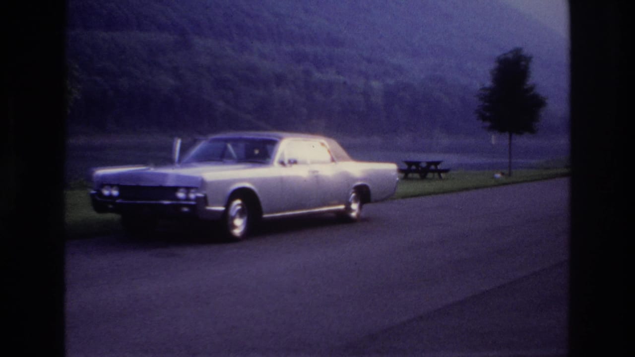 1967 Ottawa-area 8mm frame showing a silver sedan by a lakeside road, with a picnic table, trees and wooded hills behind it.