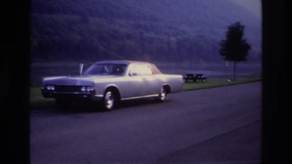 1967 Ottawa-area 8mm frame showing a silver sedan by a lakeside road, with a picnic table, trees and wooded hills behind it.
