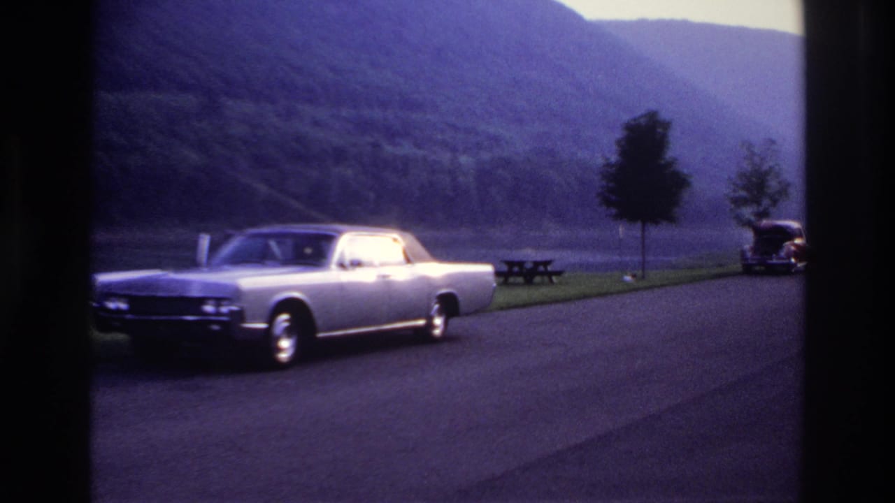 Silver sedan on a lakeside road with picnic table, trees, hills and a distant dark car.