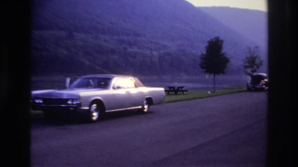 1967 Ottawa-area home-movie frame of a silver sedan beside a lake, with a picnic table, small trees, hills and a distant car visible.